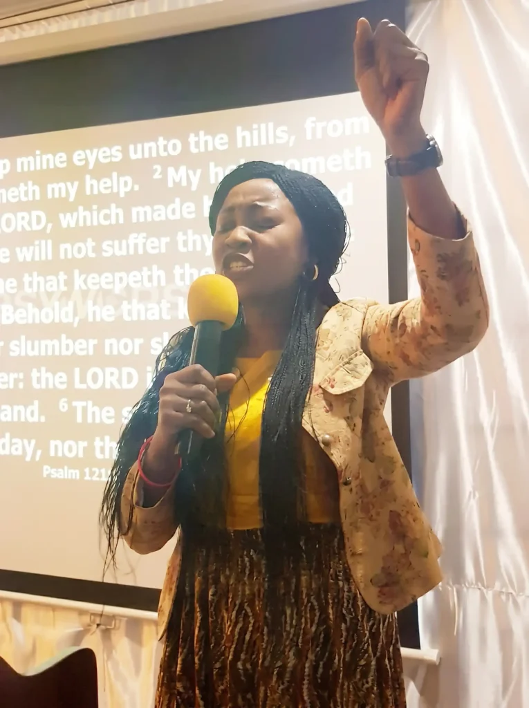 Vanessa Musenja ministering with a hand lifted, Scripture on the screen behind
